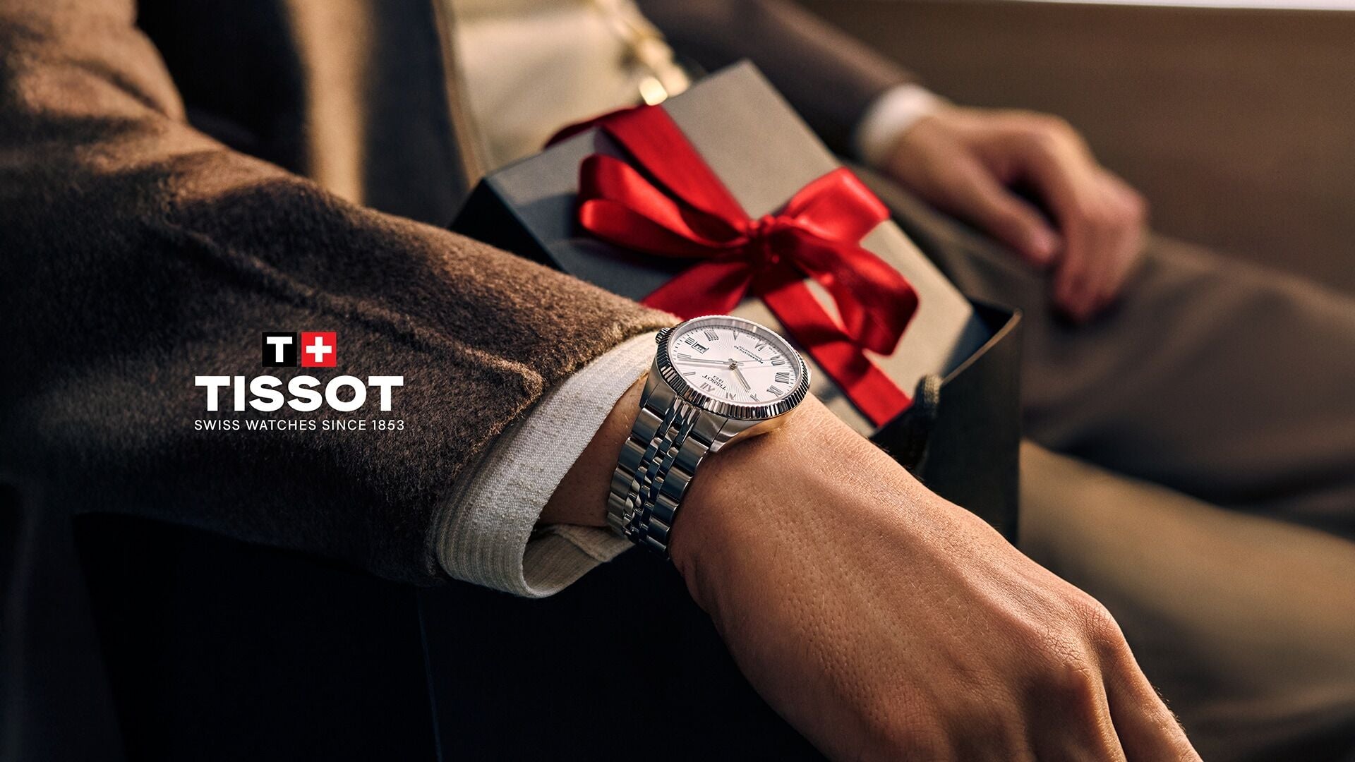 Person wearing a Tissot watch with a gift box featuring a red ribbon in the background