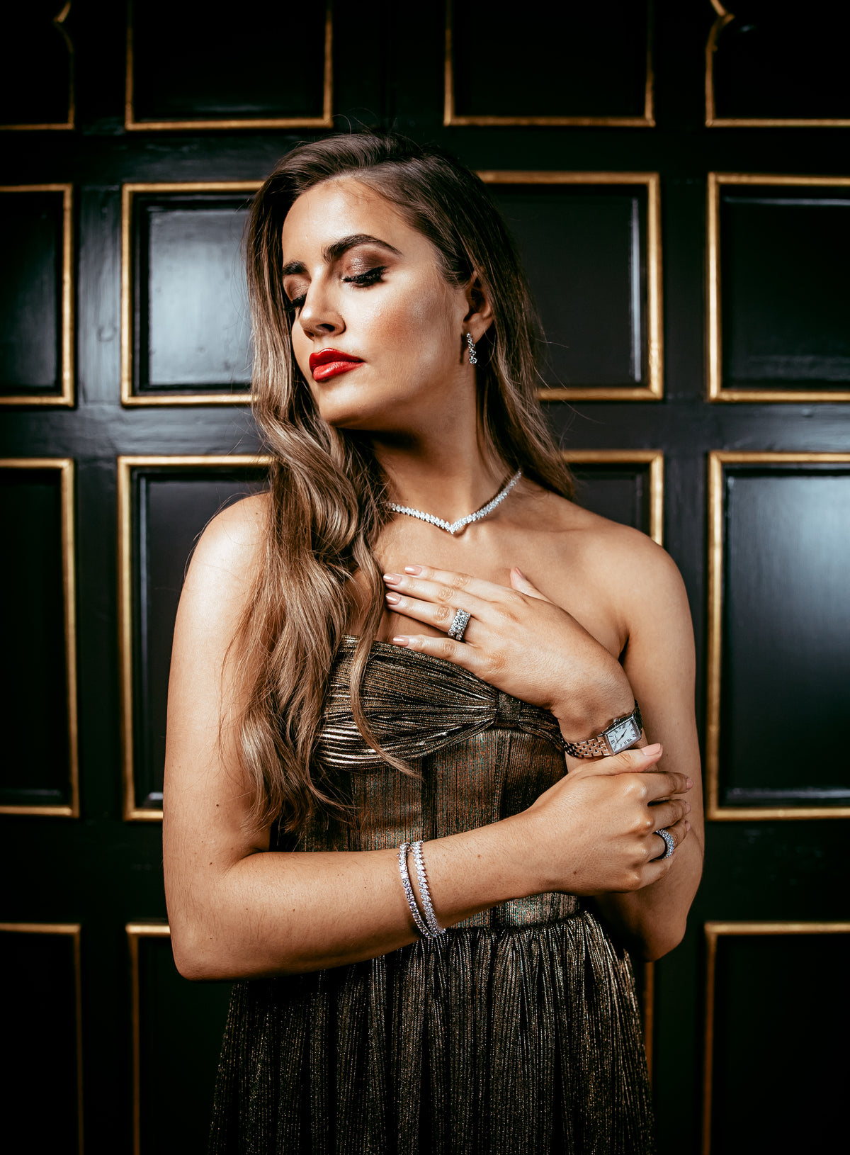 A woman with long wavy brown hair poses in a strapless metallic bronze dress, wearing a silver necklace, bracelet, and rings, set against a dark wood panel background.